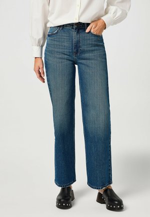 Jeans in denim blu con vita alta, gamba dritta e cuciture minime. Abbinati a zoccoli neri con borchie argentate.