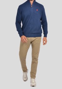 Marinblå zipkrage-tröja i mjukt tyg, ribbade manschetter och nederkant, med röd hästlogotyp. Kombinerad med beiga byxor och vita sneakers.