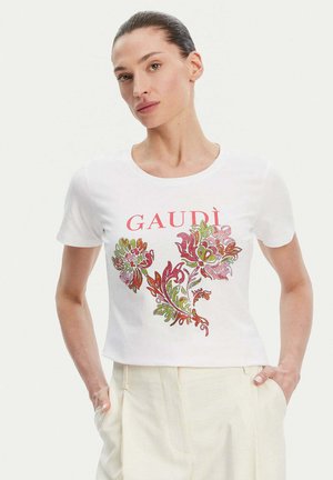 Donna che indossa una maglietta bianca con la scritta rossa "GAUDÌ" e un design floreale, abbinata a pantaloni beige chiaro a vita alta, con le mani nelle tasche.