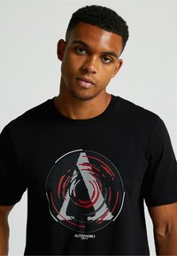 Čierne bavlnené tričko s okrúhlym grafickým dizajnom s koncentrickými čiarami v červenej a bielej farbe, so stylizovaným písmenom "A" na strede.