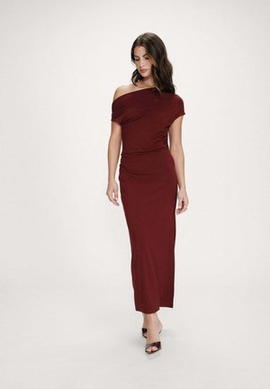 Femme marchant dans une robe bordeaux longue jusqu'au sol, épaules dénudées, avec des détails froncés, portant des talons ouverts noirs et un collier chaîne en or.