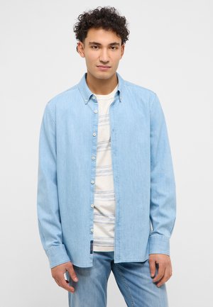 Junger Mann mit lockigem Haar, der ein hellblaues Jeanshemd über einem gestreiften weißen T-Shirt und passende Jeans trägt.