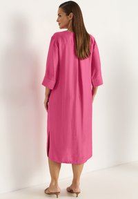 Langes, lockeres Kleid aus lebhaftem pinkem Stoff. Es hat Dreiviertelärmel, seitliche Schlitze und einen einfachen runden Ausschnitt.