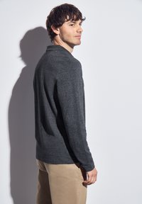 Muž s tmavými vlnitými vlasy, oblečený v tmavě šedém tričku s dlouhým rukávem a béžových kalhotách, stojí bokem proti bílé zdi.