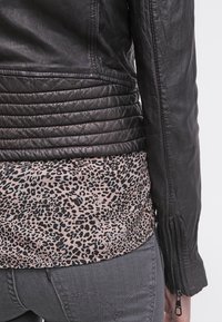 Veste en cuir noire avec matelassage horizontal sur le bas du dos, chemise à imprimé léopard en dessous, et jean en denim gris avec coutures visibles.