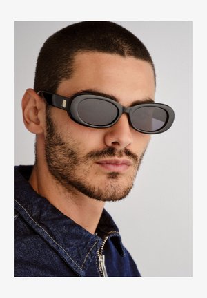 Lunettes de soleil ovales noires avec des verres foncés et une monture élégante, dotées d'un accent métallique sur les branches, portées sur un fond neutre.