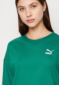 Camicetta verde a maniche lunghe in cotone, con scollatura a V e un piccolo logo bianco Puma nella parte sinistra del petto.