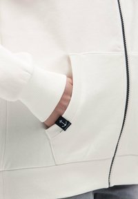 White zip-up hoodie with ribbed cuffs and a front pocket. Features a small black tag with an anchor logo on the pocket. Soft fabric texture.