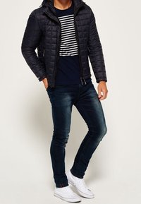 Tikkitakki hupulla, musta, vetoketjulla suljettava, puettu tummansinisen raidallisen paidan päälle. Yhdistetty tummiin farkkuihin ja valkoisiin lenkkareihin.