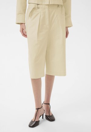 Beżowe, szerokie szorty do kolan noszone z dopasowaną bluzką oraz czarne sandały na obcasie z paskiem wokół kostki na stojącej osobie.