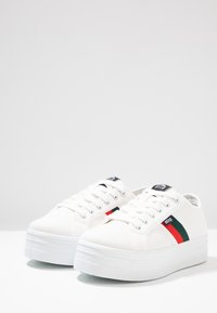 Baskets en toile blanches avec une semelle épaisse blanche, ornées d'une rayure verte et rouge et d'un accent logo noir sur la languette.