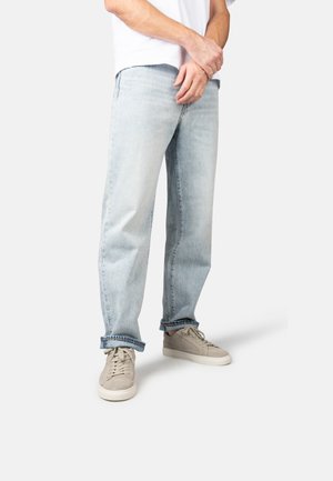 Homme portant un jean bleu clair retroussé et des baskets beiges, debout les mains jointes devant lui, sur un fond blanc uni.