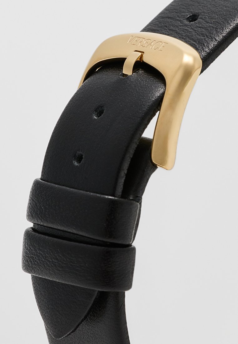 Bracelet en cuir noir avec boucle en métal doré, présentant une finition texturée et plusieurs trous pour le réglage. Détail gravé "VERSACE" sur la boucle.