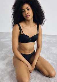 Schwarzes Dessous-Set bestehend aus einem BH mit glatten Cup-Design, verstellbaren Trägern und passendem hochgeschnittenen Bikini, alles auf einem hellen Hintergrund.