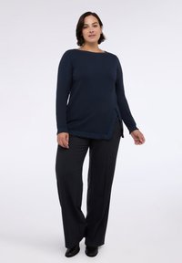 Marineblaues Langarmoberteil mit texturiertem Ausschnitt und seitlichem Schlitz, kombiniert mit dunklen weit geschnittenen Hosen und schwarzen Schuhen. Schlichtes, lässiges Design.