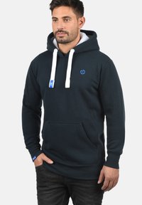 Námornícka mikina s bielymi šnúrkami, prednou klokankou, rebrovanými manžetami a malým modrým logom na hrudi. Mäkká vnútorná podšívka.