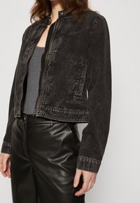 Svart jeansjacka med en kortad design, tryckknappskrage och dragkedjestängning. Har framfickor och subtilt sliten textur.