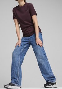 Person som bär en mörklila Puma-t-shirt och vida blå jeans, poserar med benen isär och händerna vilande på låren mot en neutral bakgrund.