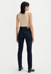 Mörkblå denimjeans med slim fit, hög midja, kontrasterande orange sömnad och en brunt läderpatch bak; svarta lägenheter. 