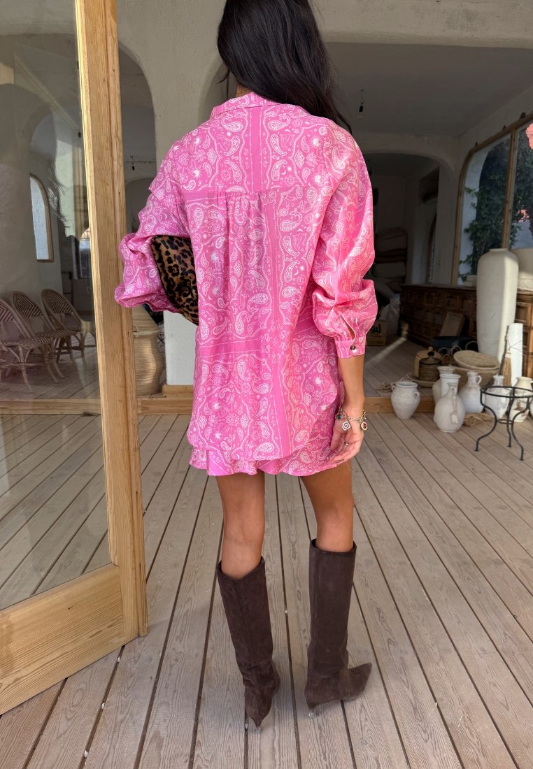 Γυναίκα με μακριά σκούρα μαλλιά σε ροζ φόρεμα με πασέ μοτίβο, κρατώντας τσάντα με σχέδιο λεοπάρδαλης, φορώντας καφέ μπότες σουέτ μέχρι το γόνατο σε εσωτερικό χώρο.