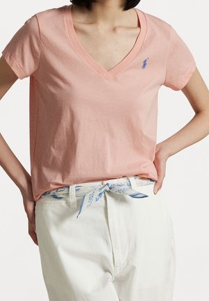 Kobieta ubrana w jasnoróżowy t-shirt z dekoltem w serek z małym niebieskim logo, białe spodnie i biały pasek z niebieskim liściastym wzorem zawiązany kokardą.