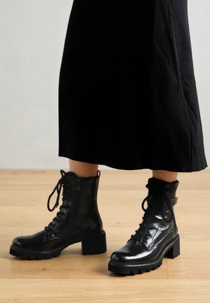 Bottines noires brillantes à lacets avec talons blocs portées avec une longue jupe noire sur un sol en bois.