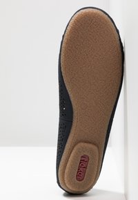 Chaussure noire avec une semelle en caoutchouc texturé et un dessus perforé. Fond en caoutchouc brun lisse, avec un détail de logo rouge près du talon.