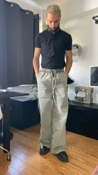 Homem com cabelo loiro encaracolado e barba, vestindo uma camisa preta e jeans cinza folgados, de pé dentro de casa com as mãos nos bolsos.