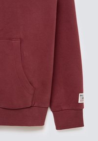 Felpa bordeaux con tasca a marsupio, polsini e orlo a coste. Presenta un'etichetta in tessuto in basso a destra con il nome del brand.