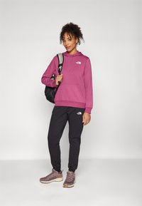 Sudadera cu hoodie din bumbac roz, prevăzută cu o buzunar frontal și un logo. Asortată cu pantaloni de trening negri și pantofi sport maro cu accente gri.