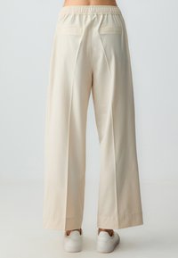 Pantaloni beige a gamba larga con una cintura elastica, dotati di due tasche posteriori e un tessuto liscio e texturizzato. Sneakers bianche visibili ai piedi.