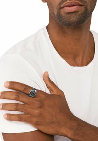 Černý kovový prsten s kruhovým designem, se zvýšeným logem ve středu. Nosen na pravé ruce, proti obyčejnému bílému tričku.