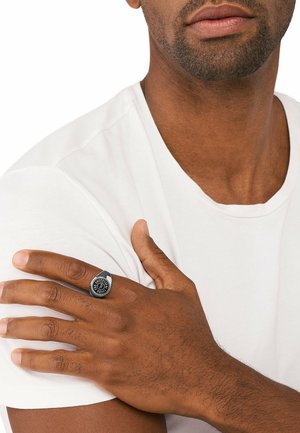 Sort metalring med et cirkulært design, der har et hævet logo i midten. Bæres på højre hånd, mod en simpel hvid T-shirt.