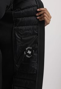 Melna izolētā jaka ar quilted iekšpusi, kura priekšpusē ir redzama kabata ar baltu logotipu un teksturētu auduma ārpusi.