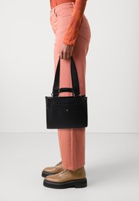 Osoba w koralowych spodniach i brązowych butach trzyma czarną torbę na dwa grube paski, stojąc na tle jasnego, jednolitego tła.