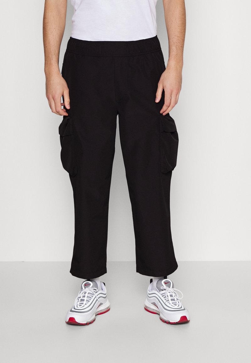 Homme portant un pantalon cargo noir avec poches latérales et des baskets blanches de sport avec des accents rouges, debout devant un fond neutre.