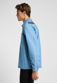Ljusblå denimjacka med avslappnad passform, som har en bröstficka och mörka accentrandar på axlarna. Knappar vid ärmsluten fulländar designen.