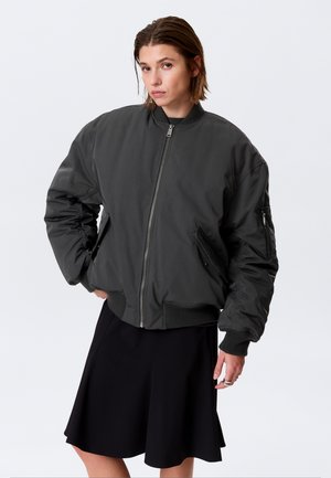 Sieviete ar īsiem matiem, valkājot tumšu oversized bomber stila jaku un melnu svārku, stāv ar vienu roku kabatā uz vienkārša fona.