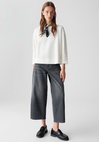 Donna che indossa un maglione bianco a trama, una sciarpa bianca e nera, jeans grigi a gamba larga e scarpe nere stringate, in piedi di fronte a uno sfondo neutro.