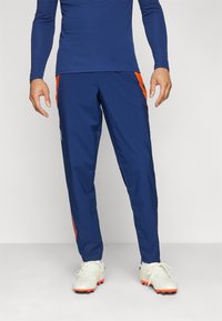 Pantaloni sportivi navy con strisce laterali arancioni, realizzati in tessuto leggero, con una vestibilità aderente e cintura elastica.