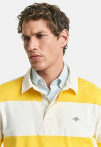 Geel en wit gestreepte polo met een geknoopte kraag, voorzien van een geborduurd logo op de borst en een gestructureerde stof.