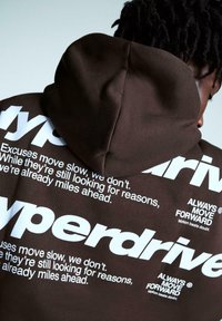 Brązowa bluza z kapturem z dużym, białym wzorem graficznym zawierającym tekst. Kaptur jest ściągany z tyłu, materiał ma gładką teksturę.