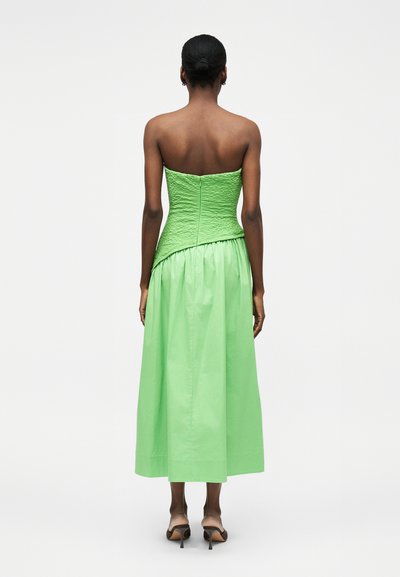 Γυναίκα με σκούρο δέρμα και μαλλιά δεμένα πίσω, ντυμένη με πράσινο τοπ χωρίς τιράντες με υφή και ταιριαστή ψηλόμεση φαρδιά φούστα, βλέποντας από πίσω.