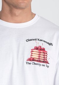 Camiseta blanca de algodón con un gráfico de pilas de pancakes cubiertas de cerezas, con el texto "Gianni Kavanagh" y "The Cherry on Top".