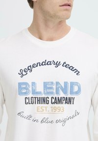 Fehér pamut póló kék és sötétkék felirattal: "Legendás csapat BLEND CLOTHING COMPANY ALAPÍTVA: 1993" egy írásos kiegészítéssel alatta.