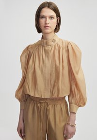 Korte mouwen, cropped top in lichtbeige stof met een gestructureerde afwerking, voorzien van een hoge nek, geplooide details en een knoopaccent.