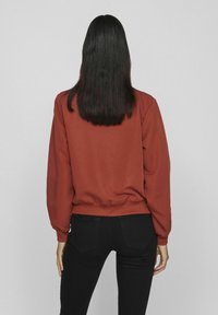 Rustfarbener Sweatshirt mit lässiger Passform, Rundhalsausschnitt und elastischem Saum. Getragen mit schwarzen Slim-Fit-Jeans, von hinten betrachtet.