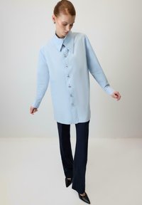 Sieviete gaiši zilā pārizmērā pogotā krekliņā, tumši zilos platā piegriezuma biksēs un melnos smailos apavos, stāv pret vienkāršu fonu.