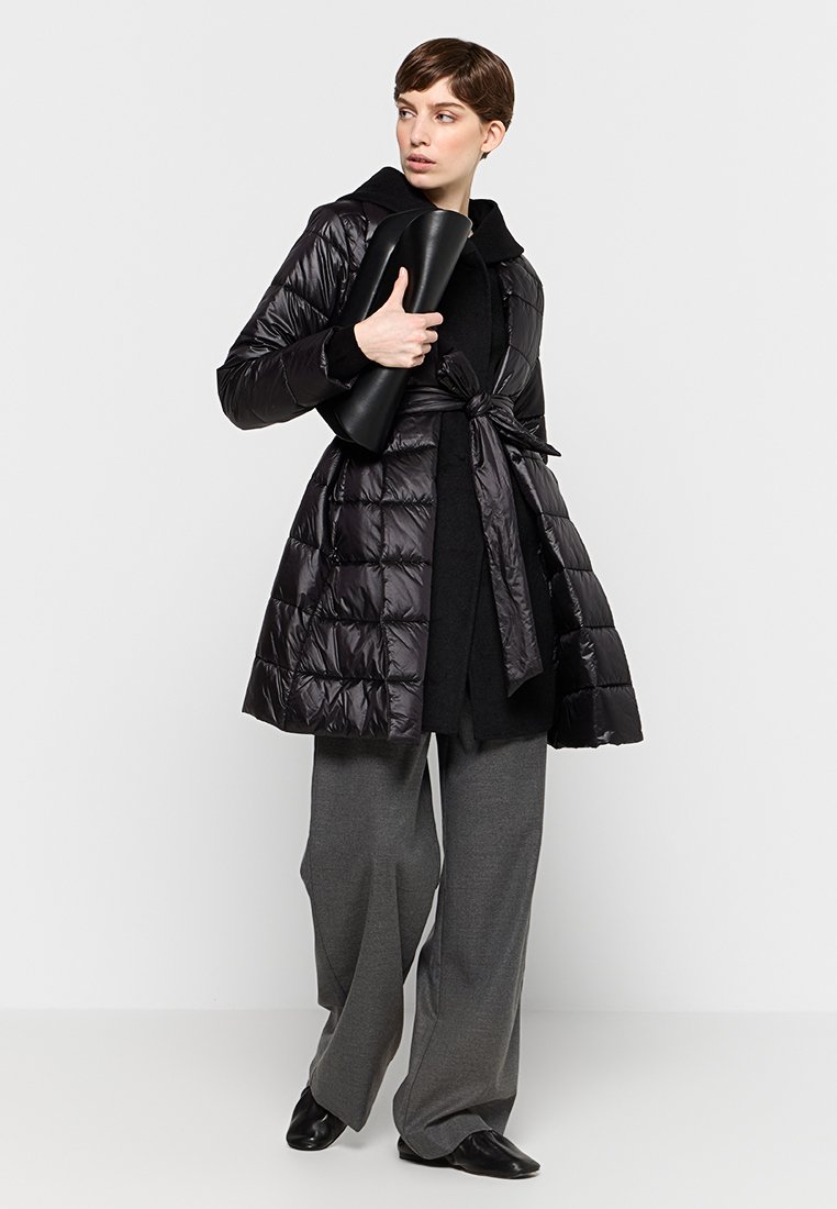 Czarna kurtka puchowa z paskiem w talii, z błyszczącym wykończeniem, dużymi kieszeniami i kołnierzem. Modelka nosi szare spodnie o szerokich nogawkach oraz czarne buty.