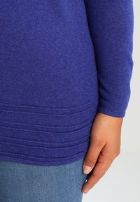 Main droite reposant le long du corps, portant un pull bleu ajusté avec un ourlet texturé et un jean bleu, sur un fond uni.